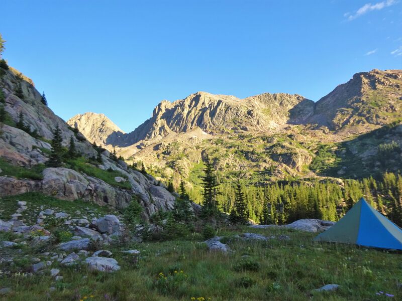 The image shows a scenic mountain landscape with a tent pitched in the foreground. The tent is blue and green, nestled in a grassy area. Towering mountains dominate the background, partially covered in trees and rocky terrain. The sky is clear and blue, suggesting a bright, sunny day. The overall scene evokes a sense of peacefulness and outdoor adventure.